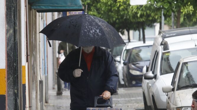 Un hombre se refugia de la lluvia bajo su paraguas a la vez que lleva mascarilla y guantes.