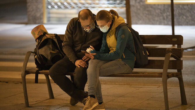 Dos personas miran el móvil sentadas en un banco.