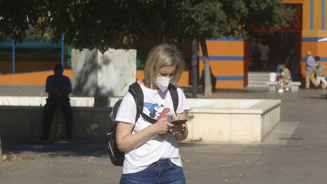 Una mujer camina protegida por su mascarilla mirando su móvil.