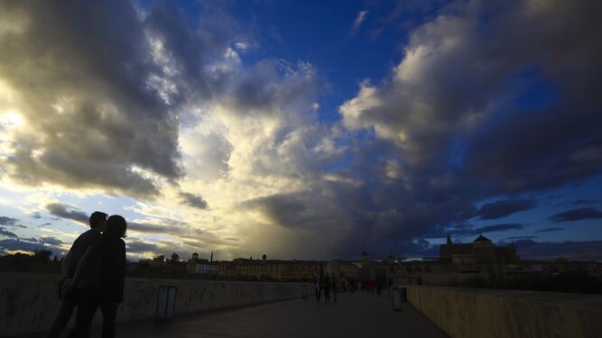 Varias personas pasean por el Puente Romano de Córdoba bajo un cielo lleno de nubes.